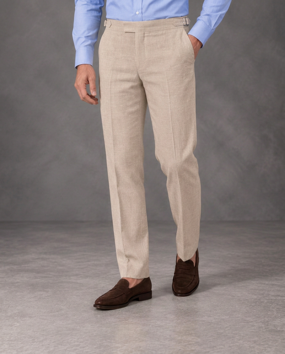 Beige straight-leg linen trousers with side adjusters, a light blue shirt and suede loafers against a grey studio backdrop